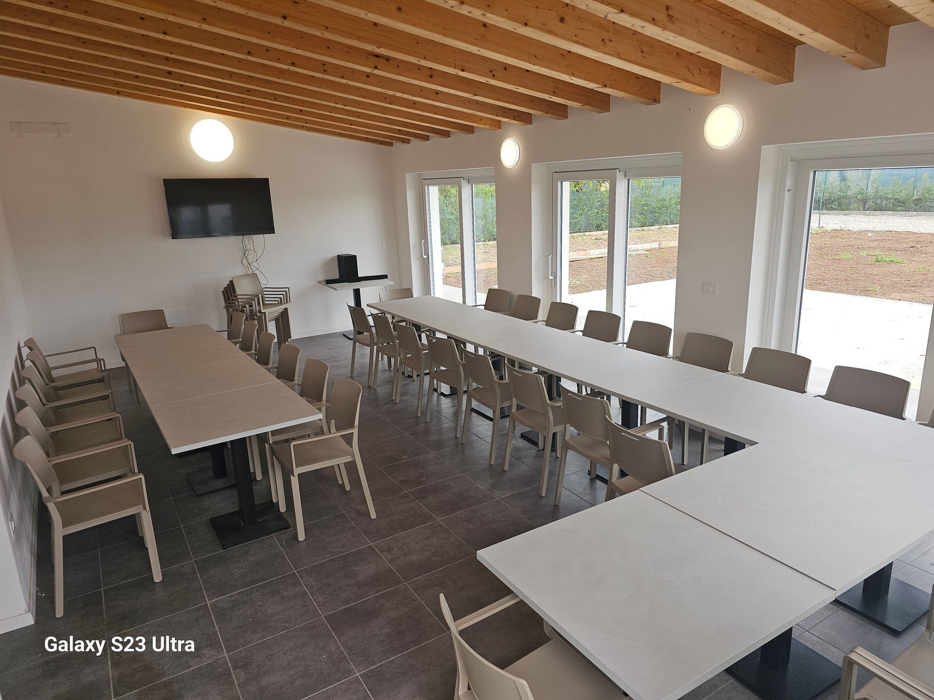 Modern meeting room with long tables, multiple chairs, wall-mounted TV, and large windows.