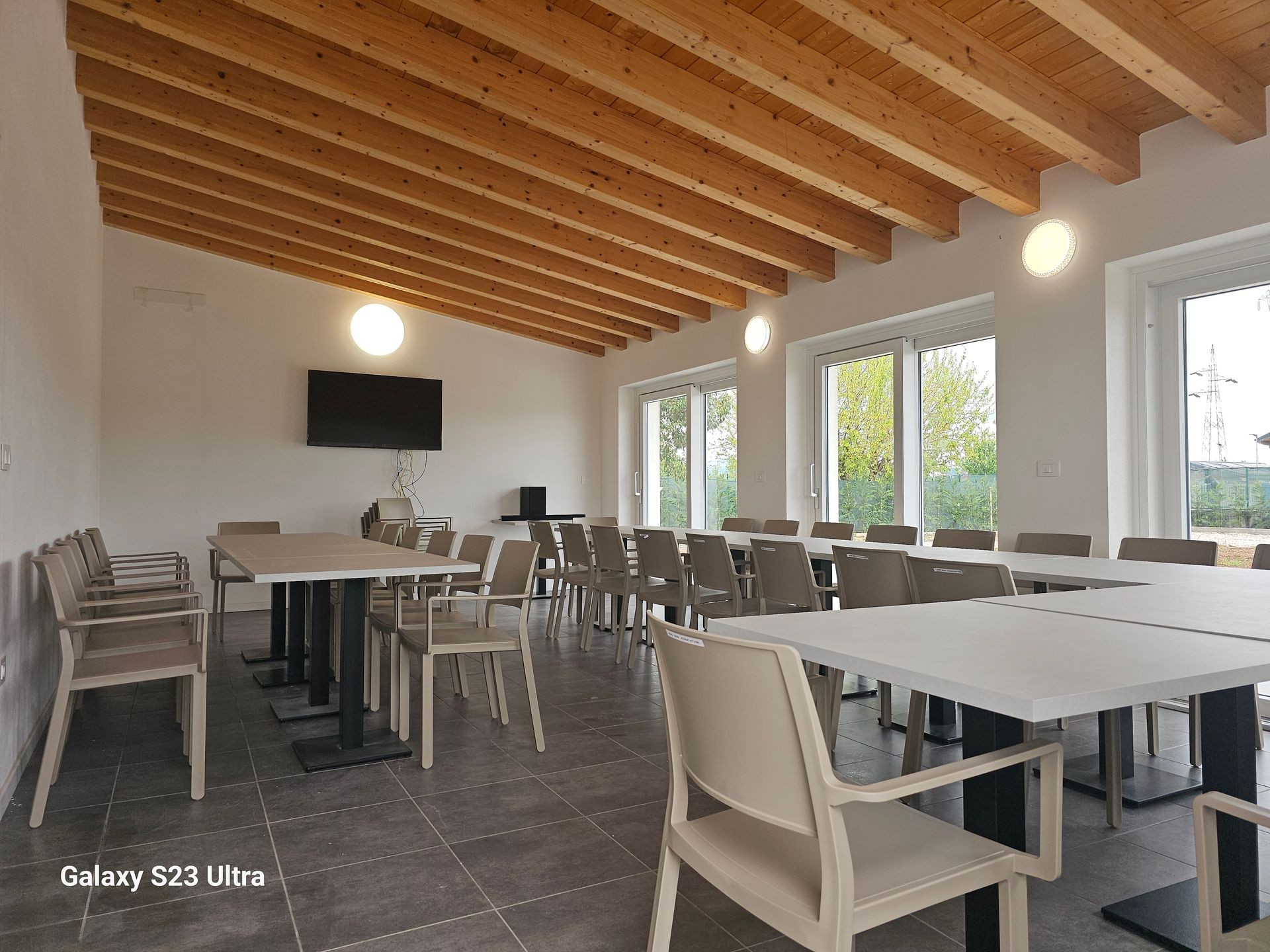 Modern conference room with wooden ceiling beams, tables, chairs, and a wall-mounted screen.